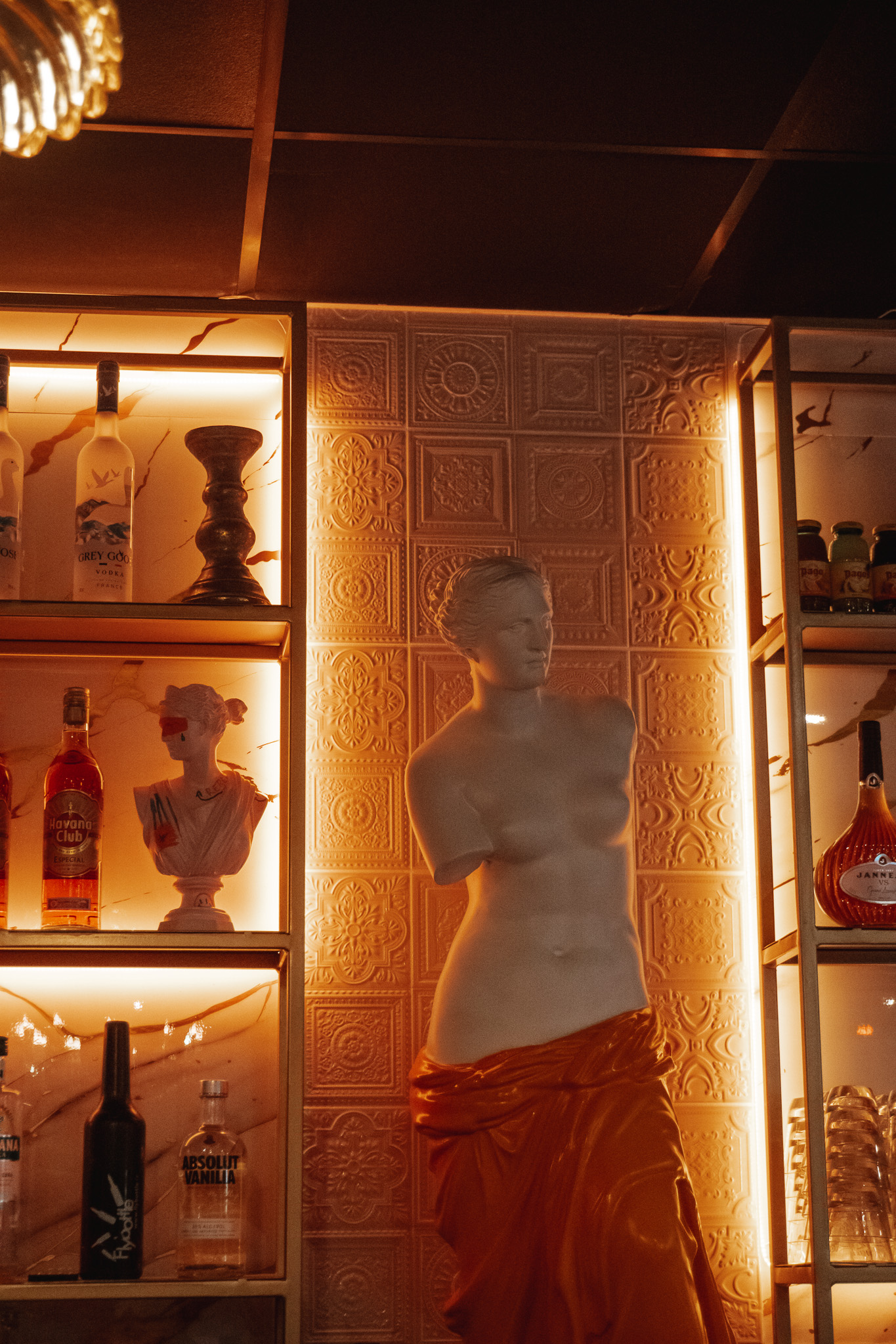 L'intérieur du bar, faiblement éclairé, présente une statue de style classique drapée d'un tissu orange, entourée d'étagères éclairées présentant des bouteilles d'alcool et des objets décoratifs sur un mur texturé.