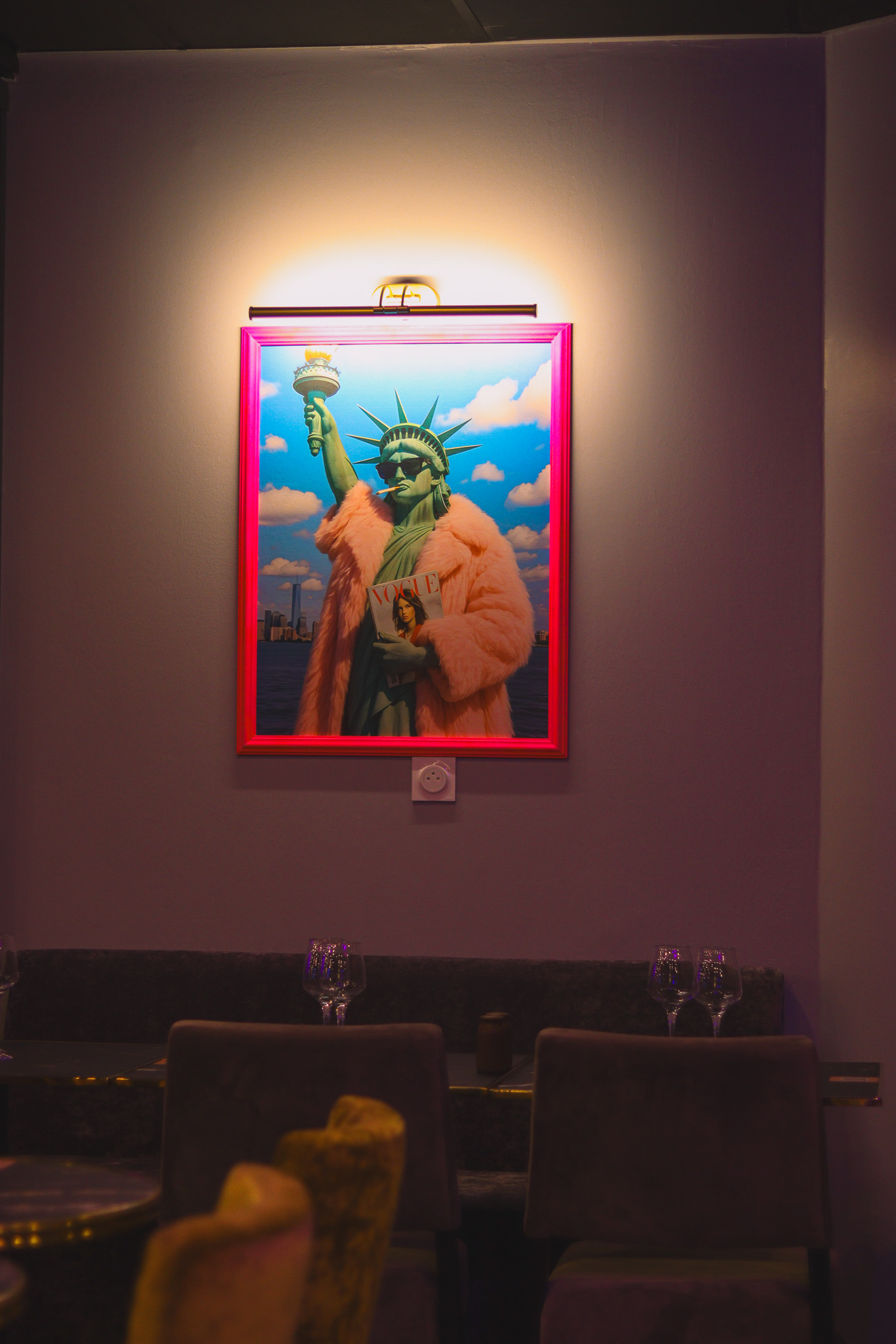 Une peinture encadrée et très lumineuse de la Statue de la Liberté portant un manteau de fourrure rose et tenant une tasse de café est accrochée à un mur gris au-dessus d'une table garnie de verres à vin vides dans une pièce faiblement éclairée.