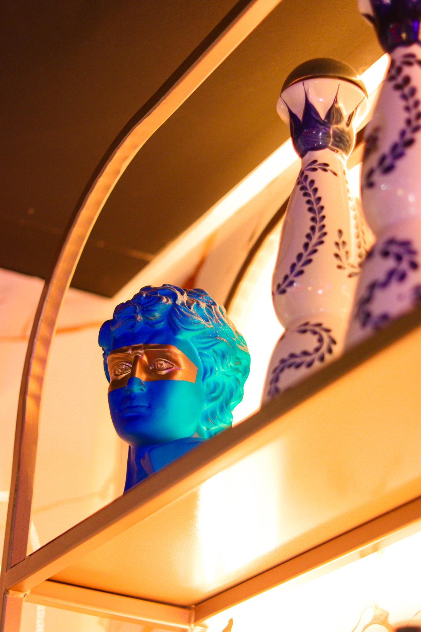 Un buste bleu avec un masque doré couvrant les yeux, exposé sur une étagère à côté d'un grand vase blanc à motifs bleus, sous un éclairage ambiant chaleureux.
