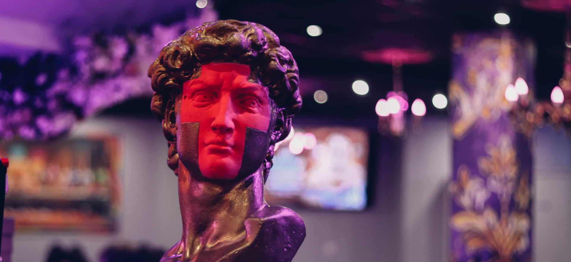 Le buste en marbre d'un homme classique est éclairé par des lumières roses et violettes, avec deux ombres rectangulaires sur son visage. L'arrière-plan est flou et présente des lustres et des décors ornés.