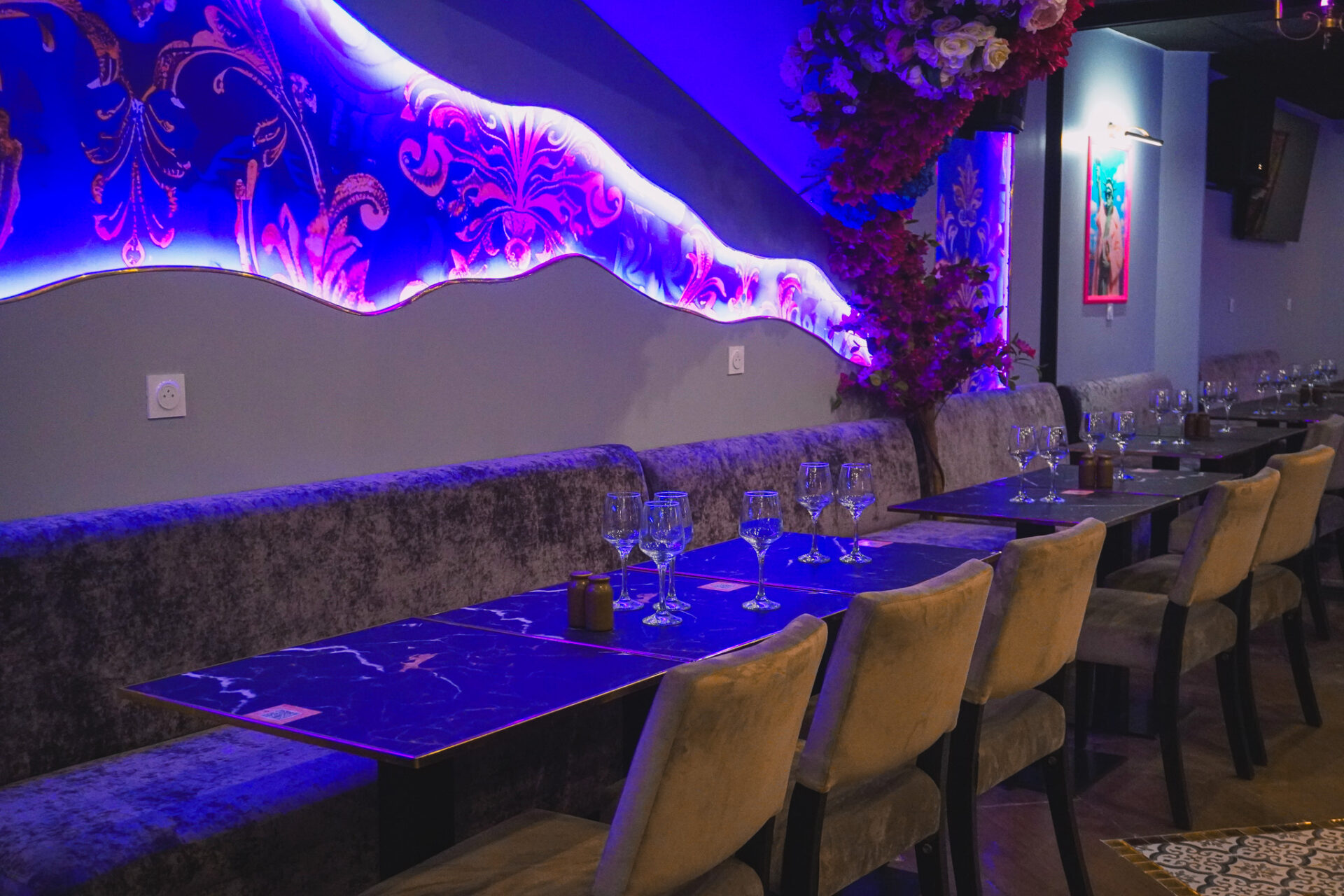 Un restaurant élégant avec des chaises en velours, des tables sombres garnies de verres et un mur ondulé décoré d'un éclairage violet et rose vibrant et d'accents floraux. L'atmosphère est moderne et accueillante.