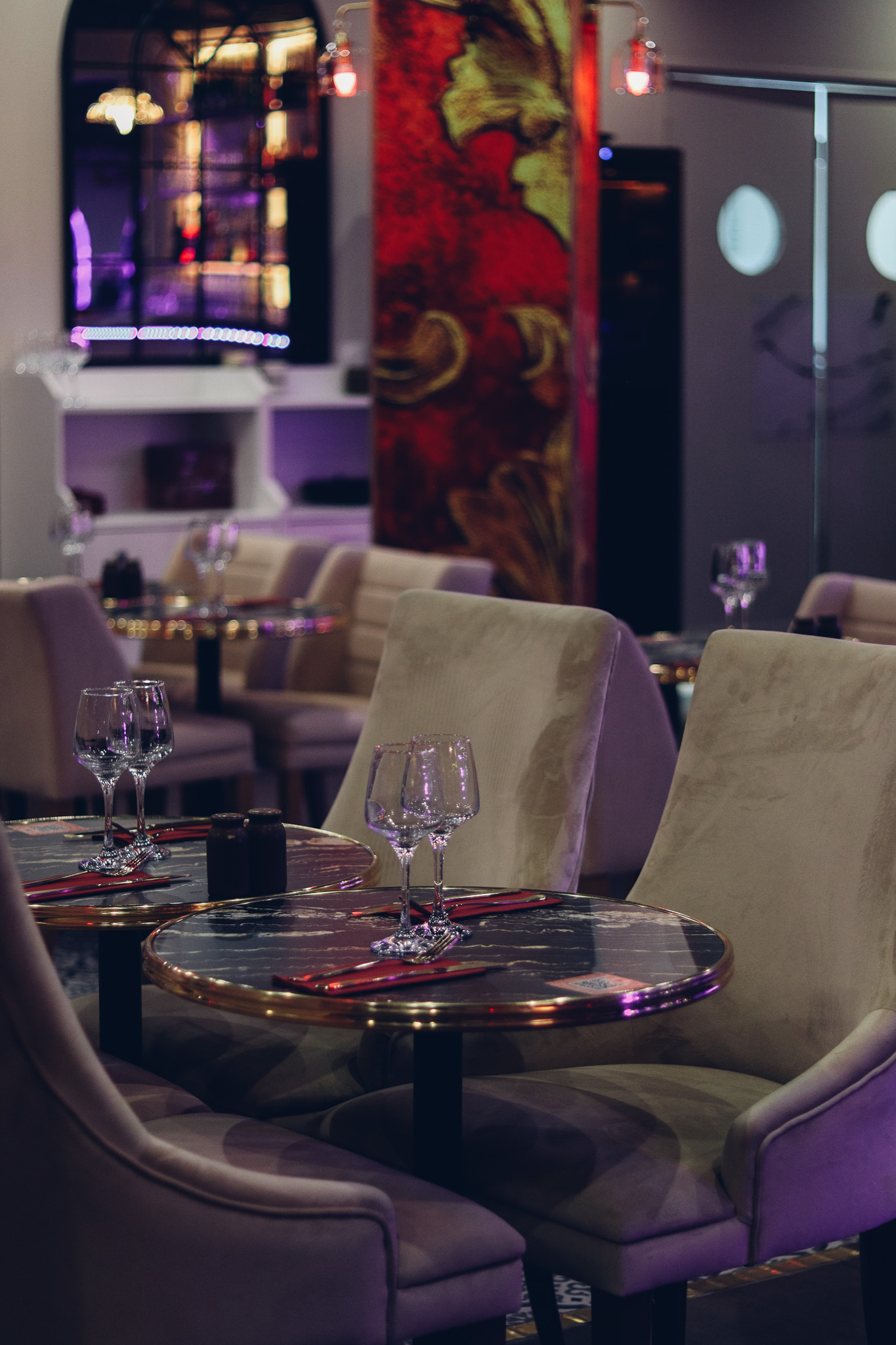 Intérieur d'un restaurant élégant avec des chaises beiges en peluche, des tables rondes en marbre garnies de verres à vin et de serviettes rouges, et un éclairage d'ambiance doux. L'arrière-plan présente un décor moderne et des œuvres d'art rouge et or éclatantes.