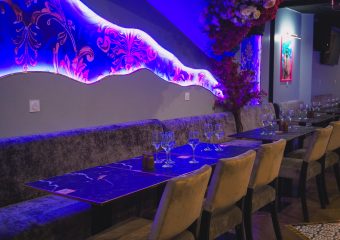 Un intérieur de restaurant moderne avec d'élégantes chaises en velours, des tables en marbre foncé garnies de verres à vin et des murs décorés de motifs floraux et de fleurs suspendues, éclairés par des diodes électroluminescentes violettes.