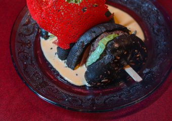 Un hamburger noir gourmet sur une assiette en verre transparent, garni de laitue verte, d'une sauce crémeuse et surmonté d'un croustillant rouge décoratif en dentelle, le tout posé sur une riche nappe rouge.